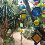 A tropical garden with plants and palm trees. There are multicoloured glass sculptures hanging down, with circular and diamond shapes.