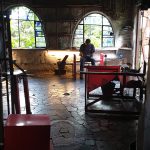 An image of the glassworks interior. It is dark and shadowy.There is a man to the rear hard at work making a glass object which we can't see.