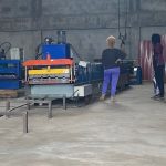 Image of factory with some people standing around. There is a long machine that has rollers on for creating corrugated roof panels from sheet metal