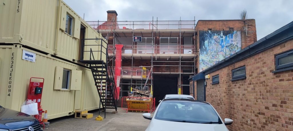 Image of a building being refurbished. It is covered in scaffolding and there are two container units in the yard in front of it, along with a full building skip.