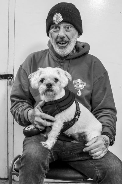 Image of man with a white beard and a wooly hat. He's sitting down with a small white dog on his lap.